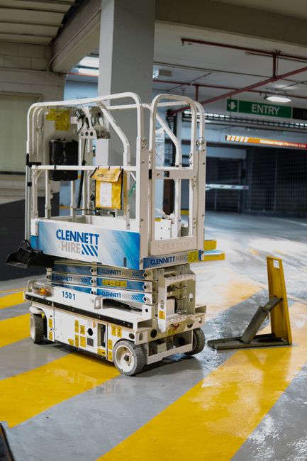 The image shows an indoor parking garage with a white mobile elevated work platform branded 'Clennett Hire' parked on a yellow and grey striped floor. The platform has a safety rail and a control pane